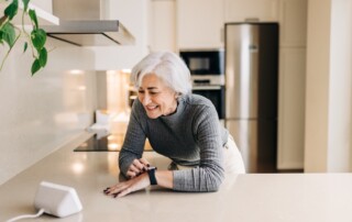 A senior woman stands in her kitchen and uses her digital assistant