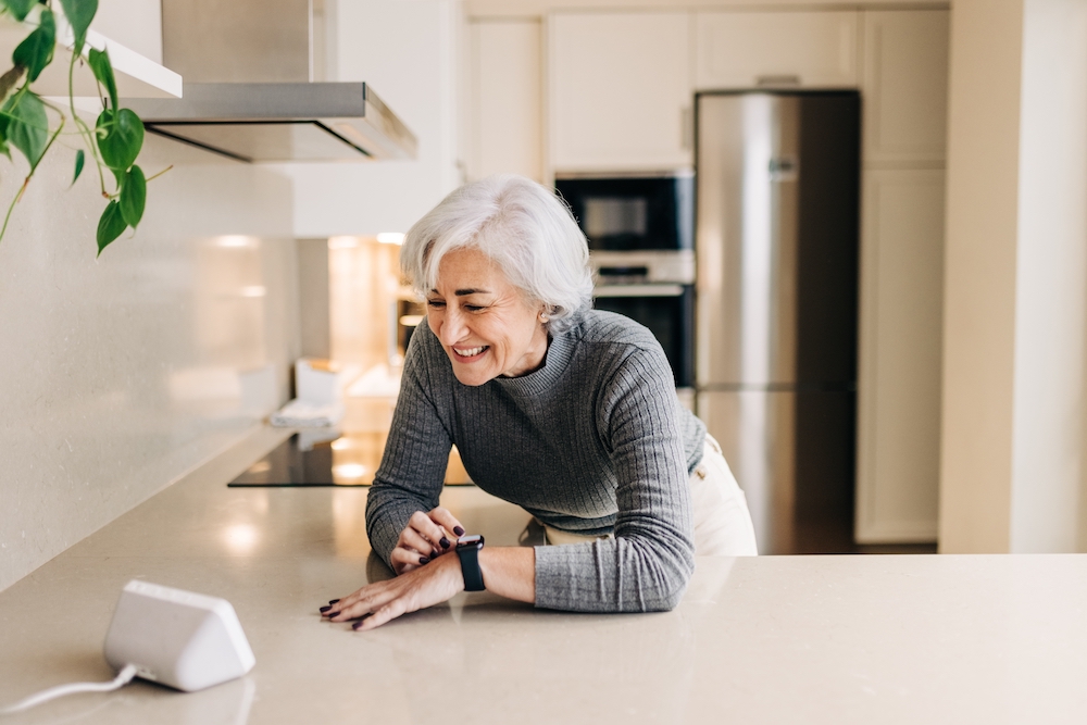 A senior woman stands in her kitchen and uses her digital assistant
